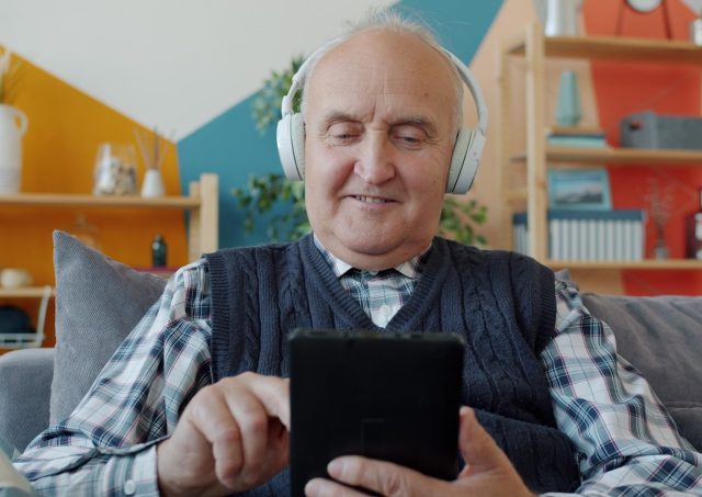 Elderly man with headphones uses a tablet on couch.